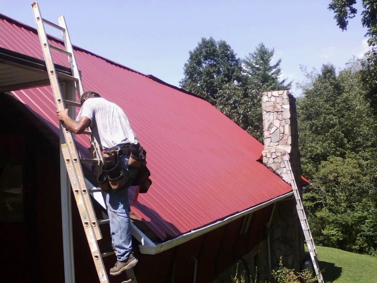 Roofer on a ladder beside a red metal roof and stone chimney — Soffit & Fascia Repair by Roofing Brothers in Lucas